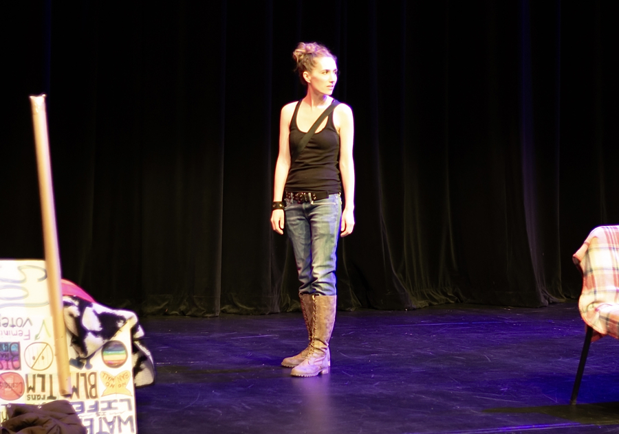 An actor is on stage looking over their shoulder, protest signs are stacked upside against the wall.