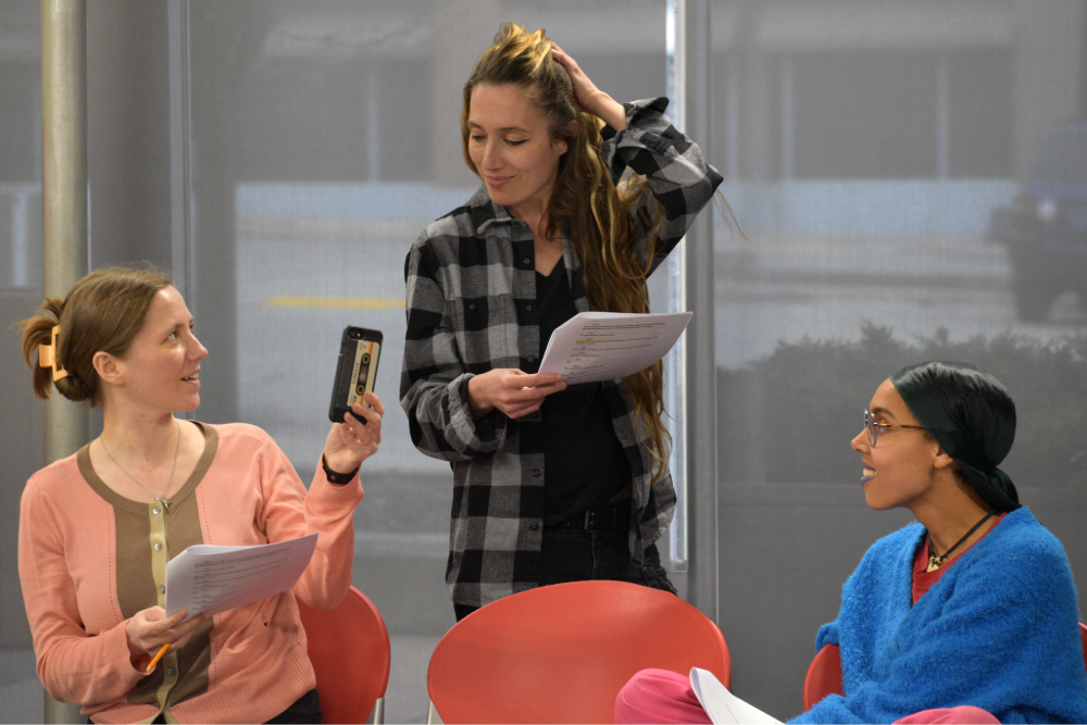 Ashely Bagot, Cyra K. Polizzi, and stella Akua performing with scripts in hand. Two of them are sitting, the other stands behind, they are smiling.